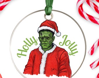 Puede incluir: Adorno navideño redondo con el monstruo de Frankenstein verde con gorro de Papá Noel y abrigo rojo. Las palabras "Holly Jolly" están escritas en verde. El adorno cuelga de una cinta verde. Bastones de caramelo rojos y blancos en el fondo.