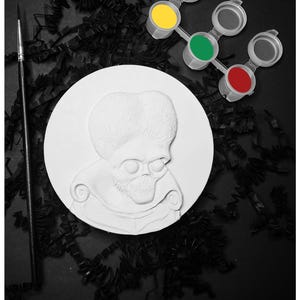 May include: A white circular plaster relief featuring a sculpted alien figure with an oversized brain and skeletal facial features, accompanied by a thin black paintbrush and a small plastic strip containing yellow, green, and red paint pots.
