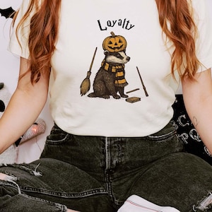May include: A cream-colored t-shirt featuring a graphic of a badger wearing a pumpkin on its head and a striped scarf, accompanied by a broom and a wand. The word "Loyalty" is printed in black script above the badger illustration.