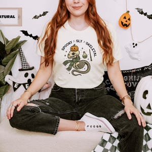 May include: A cream-colored short-sleeve t-shirt featuring a graphic of a snake skeleton wearing a green scarf, topped with a small orange pumpkin. The text "SPOOKY SLY SKELETAL" is printed in black above the illustration, which also includes a potion bottle and a wand.
