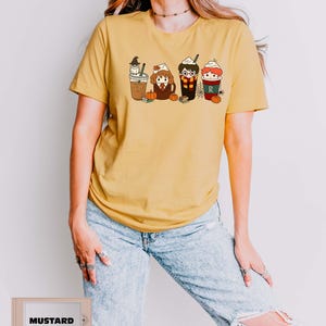 May include: Mustard yellow t-shirt featuring a cartoon graphic of four themed coffee cups. Each cup has a different character design, with whipped cream and a straw. The shirt is paired with light blue ripped jeans.