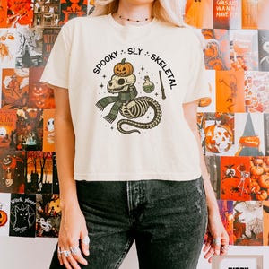 May include: An ivory short-sleeve t-shirt featuring a graphic print of a skull wearing a pumpkin on its head, wrapped in a green and black striped scarf, with a potion bottle and wand. The text reads SPOOKY SLY SKELETAL in black lettering.