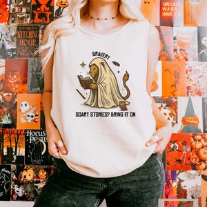 Könnte beinhalten: Ein ärmelloses elfenbeinfarbenes Tanktop mit einer grafischen Illustration eines Löwen, der ein Geisterkostüm trägt und ein Buch liest. Der Text über dem Löwen lautet BRAVERY und der Text darunter lautet SCARY STORIES? BRING IT ON. Eine kleine Eule sitzt daneben.