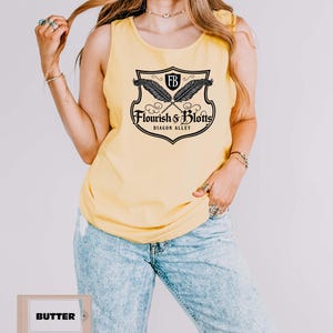 May include: Yellow tank top with a black and white graphic featuring crossed quills, a shield, and the text "Flourish & Blotts, Diagon Alley." The word "Butter" is printed on a small rectangular sign.