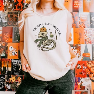 May include: An ivory sleeveless tank top featuring a graphic print of a skeleton creature wearing a green plaid scarf with a pumpkin on its head, a potion bottle, and a wand. The text "SPOOKY SLY SKELETAL" is printed in an arc above the graphic.
