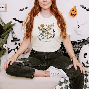 May include: A cream-coloured t-shirt featuring a graphic of a bird skeleton wearing a scarf and balancing a pumpkin on its head. The design includes a small broom and a wand, with the text "SPOOKY SMART SKELETAL" printed in a circular arrangement.