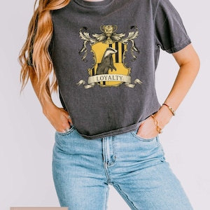 May include: Dark gray t-shirt featuring a crest with a badger, yellow and black stripes, and the word "LOYALTY" on a banner. The shirt is paired with blue jeans.