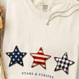May include: Cream-colored t-shirt with three star patches in navy with white stars, red and white stripes, and black and white gingham. The text "STARS & STRIPES" is printed below the stars. A silver star necklace is draped on the shirt.