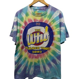 Op de afbeelding: Tie-dye T-shirt met een cirkelvormige afbeelding met de tekst "The Wind Summer Tour 2002" in geel en rood. De afbeelding bevat een Union Jack-vlag. Het shirt heeft een spiraalpatroon in blauw, paars, geel en roze.