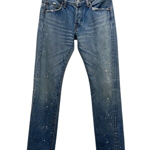 Könnte beinhalten: Eine blaue Jeans aus Denim mit abgenutztem Finish, leichter Verwaschung, Sitzfalten an den Hüften und verstreuten weißen Farbspritzern. Die Jeans verfügt über ein klassisches Fünf-Taschen-Design, einen Knopfverschluss und kupferfarbene Nieten.