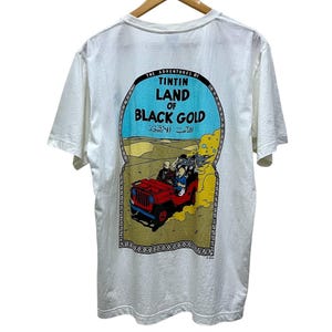 May include: White t-shirt featuring a colorful graphic from "The Adventures of Tintin: Land of Black Gold." The design includes a red jeep with characters, set against a desert backdrop, and the title text in English and Arabic.