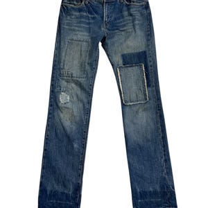 May include: Blue denim jeans with a faded wash and multiple patches on the front. The jeans have a classic five-pocket design and a button-and-zipper closure. The patches vary in size and texture, adding a distressed look.