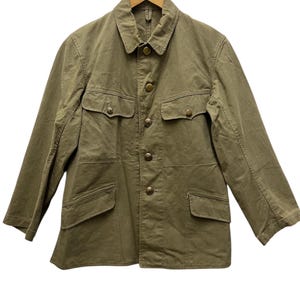 May include: Olive green jacket with four front pockets and button closures. The jacket has a collared neckline and long sleeves. It is hanging on a wooden hanger against a white background. The jacket appears to be made of a durable fabric.