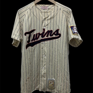 May include: Cream-colored baseball jersey with navy blue pinstripes and the word "Twins" in maroon and navy blue across the chest. Features a team logo patch on the sleeve and a small tag at the bottom. The jersey is button-down and has short sleeves.