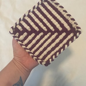 Puede incluir: Un salvamanteles cuadrado, hecho a mano, con un patrón de rayas crema y morado oscuro. Los bordes están trenzados con un cordón morado a juego, y se adjunta un pequeño lazo para colgarlo. Este accesorio de cocina está diseñado para proteger las superficies del calor.