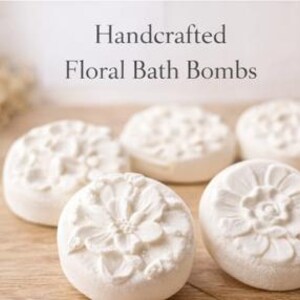 May include: Five white, floral-designed bath bombs are arranged on a wooden surface. The bath bombs have a circular shape with detailed flower patterns. The text "Handcrafted Floral Bath Bombs" is displayed above the bath bombs.