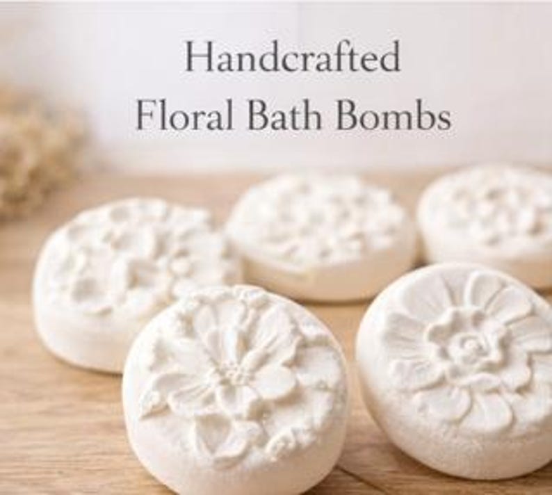May include: Five white, floral-designed bath bombs are arranged on a wooden surface. The bath bombs have a circular shape with detailed flower patterns. The text "Handcrafted Floral Bath Bombs" is displayed above the bath bombs.
