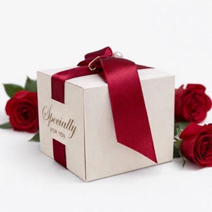 May include: A cream-colored gift box tied with a deep red satin ribbon and a pearl accent. The box has the words "Specially For You" in gold script. Red roses are placed around the box.