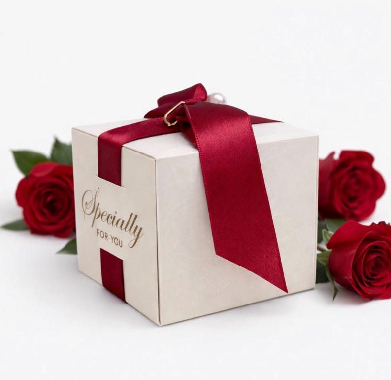 May include: A cream-colored gift box tied with a deep red satin ribbon and a pearl accent. The box has the words "Specially For You" in gold script. Red roses are placed around the box.