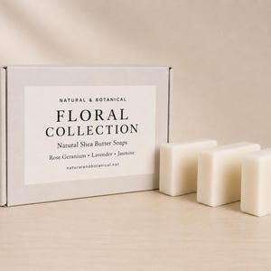 May include: A light grey rectangular cardboard box labelled NATURAL & BOTANICAL FLORAL COLLECTION Natural Shea Butter Soaps Rose Geranium Lavender Jasmine naturalandbotanical.net with three off-white rectangular soap bars and folded towels.