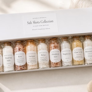 May include: A white rectangular gift box containing ten small glass bottles with cork stoppers, each filled with different coloured bath salts. The label on the box reads Salt Shots Collection. Each bottle has a white oval label with text.