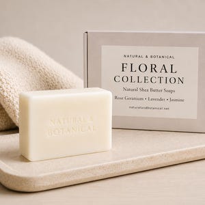 May include: A rectangular off-white bar of Natural & Botanical shea butter soap embossed with text, positioned next to a matching cardboard box labelled Floral Collection with scents Rose Geranium, Lavender, and Jasmine, and a beige knit fabric.