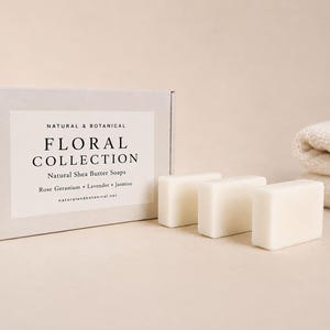 May include: A rectangular light grey cardboard box labelled NATURAL & BOTANICAL FLORAL COLLECTION Natural Shea Butter Soaps Rose Geranium Lavender Jasmine naturalandbotanical.net with three solid white rectangular soap bars and a folded cloth.