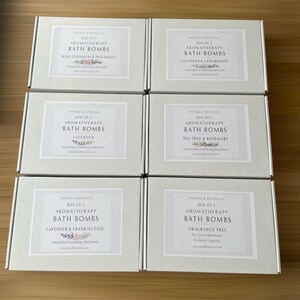 May include: Six boxes of aromatherapy bath bombs are displayed, each with a different scent: Rose Geranium & Patchouli, Lavender & Cedarwood, Lavender, Tea Tree & Rosemary, Lavender & Frankincense, and Fragrance Free. The boxes are light green with white labels.