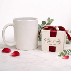 May include: A white ceramic mug with a curved handle sits next to a small gift box tied with a burgundy ribbon and a pearl accent. The box reads "Specially FOR YOU" in gold lettering. Red rose petals are scattered around.