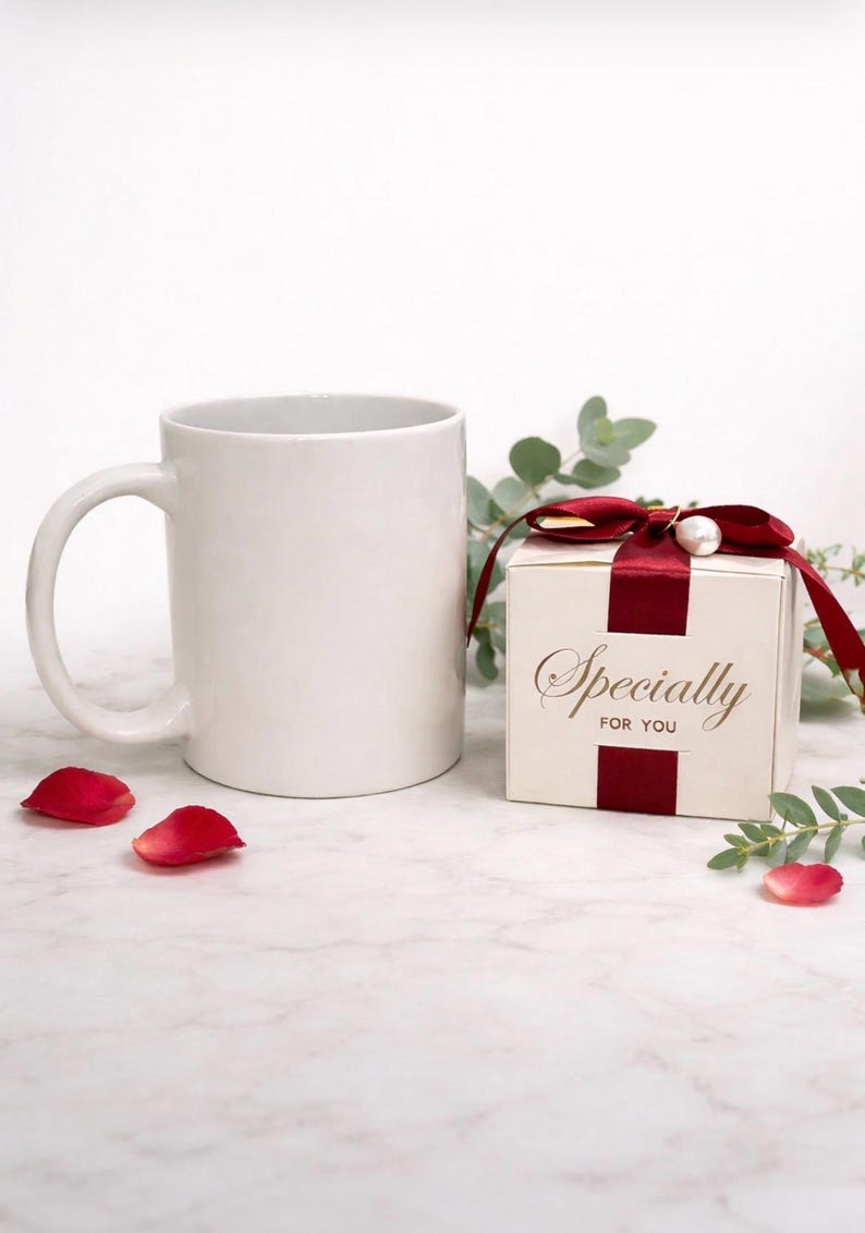 May include: A white ceramic mug with a curved handle sits next to a small gift box tied with a burgundy ribbon and a pearl accent. The box reads "Specially FOR YOU" in gold lettering. Red rose petals are scattered around.