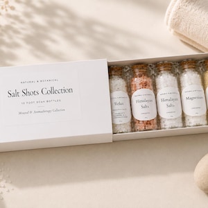 May include: A white sliding gift box labelled Salt Shots Collection containing five small glass bottles filled with bath salts. The labels read Relax, Himalayan Salts, Himalayan Salts, Magnesium, and Glow 'n' Go, each with 15g net weight.