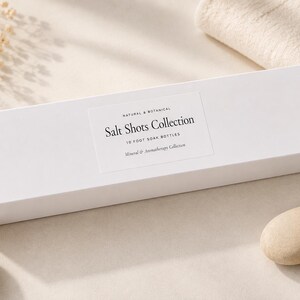 May include: A rectangular white box labelled Salt Shots Collection 10 Foot Soak Bottles Mineral & Aromatherapy Collection. The packaging features black serif text on a white label centred on the box, presenting a clean and minimalist aesthetic.