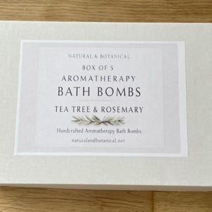 May include: A white box with a label that reads "NATURAL & BOTANICAL BOX OF 5 AROMATHERAPY BATH BOMBS TEA TREE & ROSEMARY." The label also says "Handcrafted Aromatherapy Bath Bombs" and "naturalandbotanical.net."