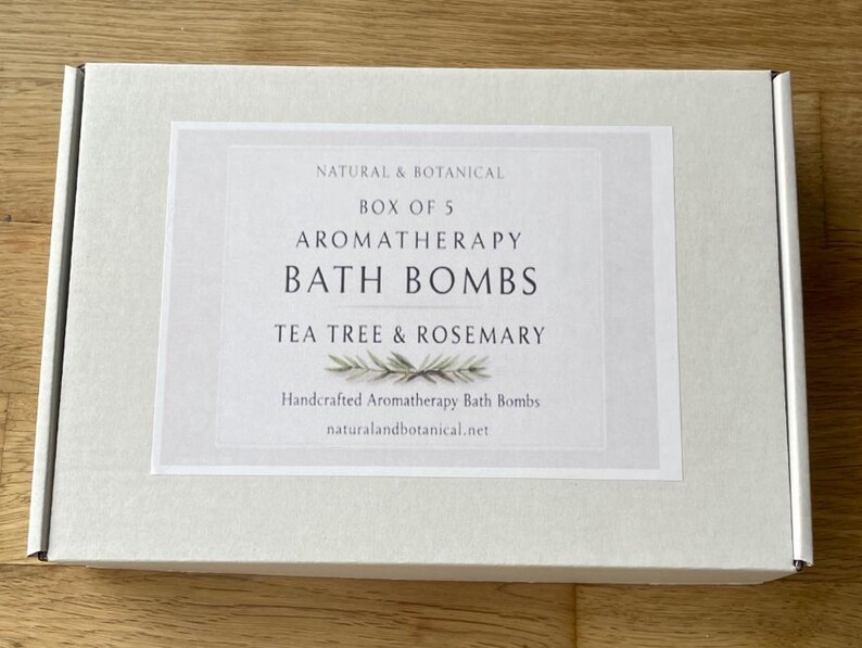 May include: A white box with a label that reads "NATURAL & BOTANICAL BOX OF 5 AROMATHERAPY BATH BOMBS TEA TREE & ROSEMARY." The label also says "Handcrafted Aromatherapy Bath Bombs" and "naturalandbotanical.net."