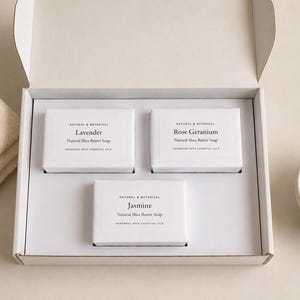 May include: A white gift box containing three rectangular bars of natural shea butter soap wrapped in white paper. Each label features the text Natural & Botanical, the scent name Lavender, Rose Geranium, or Jasmine, and Handmade with Essential Oils.