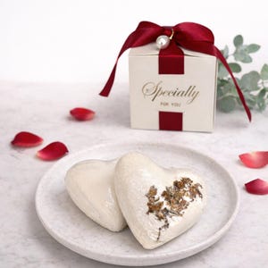 May include: Two heart-shaped, white bath bombs with gold accents rest on a speckled plate. A small gift box with a red ribbon and the words "Specially For You" is in the background. Red rose petals are scattered around the plate.