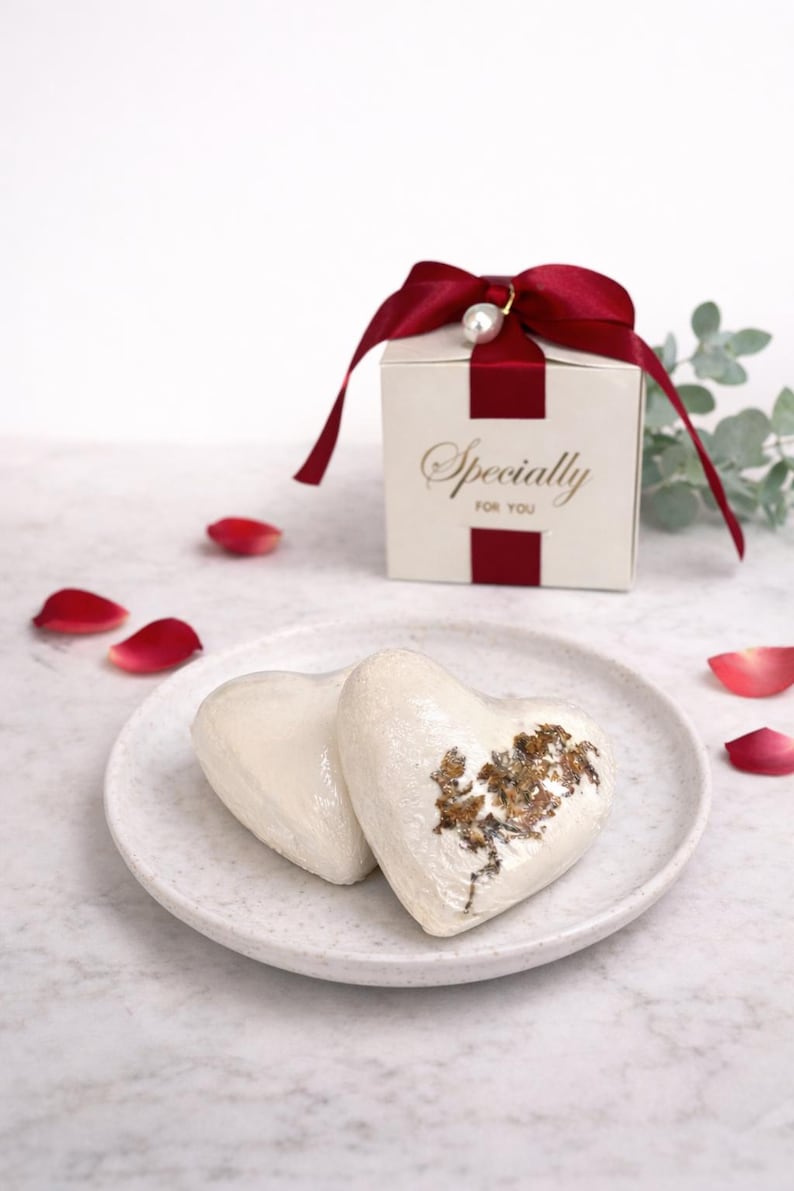 May include: Two heart-shaped, white bath bombs with gold accents rest on a speckled plate. A small gift box with a red ribbon and the words "Specially For You" is in the background. Red rose petals are scattered around the plate.