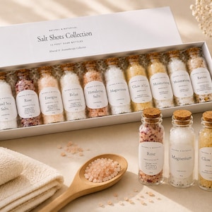 May include: A Salt Shots Collection set featuring twelve small glass bottles filled with various bath salts, each with a cork stopper and white label. Three additional bottles sit in front, alongside a wooden spoon filled with pink salt crystals.
