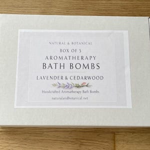 May include: A white box with a label that reads "NATURAL & BOTANICAL BOX OF 5 AROMATHERAPY BATH BOMBS LAVENDER & CEDARWOOD." The label also includes the text "Handcrafted Aromatherapy Bath Bombs" and the website address.