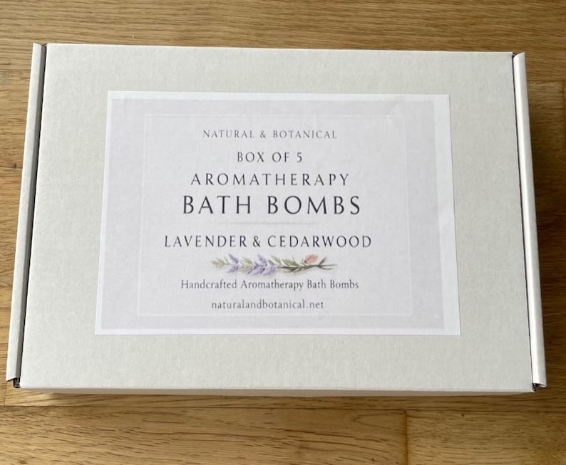 May include: A white box with a label that reads "NATURAL & BOTANICAL BOX OF 5 AROMATHERAPY BATH BOMBS LAVENDER & CEDARWOOD." The label also includes the text "Handcrafted Aromatherapy Bath Bombs" and the website address.