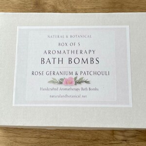 May include: A white box with a label that reads "NATURAL & BOTANICAL" and "AROMATHERAPY BATH BOMBS." The label also states "ROSE GERANIUM & PATCHOULI" and "Handcrafted Aromatherapy Bath Bombs." The box is on a wooden surface.