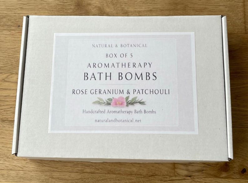 May include: A white box with a label that reads "NATURAL & BOTANICAL" and "AROMATHERAPY BATH BOMBS." The label also states "ROSE GERANIUM & PATCHOULI" and "Handcrafted Aromatherapy Bath Bombs." The box is on a wooden surface.