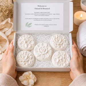 May include: A white box filled with five round, floral-designed soaps. The soaps are arranged on white paper filler. The box has a printed message that reads "Welcome to Natural & Botanical". A lit candle and a rolled towel are in the background.