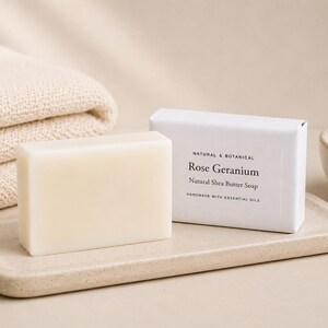 May include: A rectangular bar of off-white soap next to its white paper packaging. The packaging features the text: NATURAL & BOTANICAL, Rose Geranium, Natural Shea Butter Soap, HANDMADE WITH ESSENTIAL OILS. The soap rests on a beige tray.