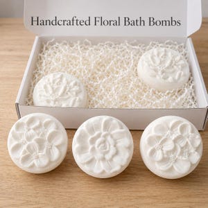 May include: A white box filled with white shredded paper, containing six white, floral-designed bath bombs. The bath bombs have different embossed floral patterns. The box reads "Handcrafted Floral Bath Bombs".