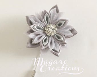 May include: A gray fabric flower brooch with a silver center and a sparkling rhinestone embellishment. The flower has multiple petals and a long pin backing. The text "Magaro Creations" is in the lower right corner.