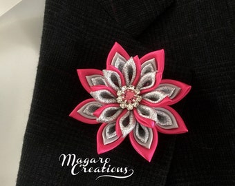 May include: A pink and silver flower brooch with a sparkly rhinestone center. The brooch is pinned to a black fabric background.