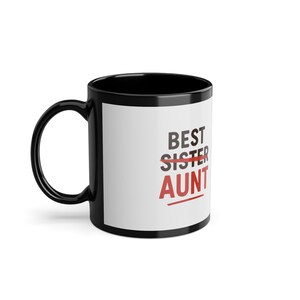 Può includere: Tazza da caffè nera con interno bianco e manico nero. La tazza presenta la scritta "BEST AUNT" in nero e rosso, con "SISTER" cancellato. Un regalo per una zia.
