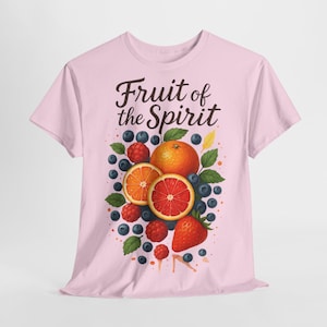 Può includere: T-shirt rosa chiaro con un'illustrazione di frutta e la scritta "Fruit of the Spirit". Il design include arance, fragole, mirtilli e lamponi. La maglietta è realizzata in materiale morbido.