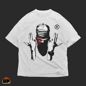 Könnte beinhalten: Weißes T-Shirt mit einer schwarz-weißen Grafik einer Person, die eine Kopfbedeckung und ein Ziel über dem Gesicht trägt. Das Design beinhaltet zwei Hände, die eine Handgeste machen. Ein eingetragenes Warenzeichen ist sichtbar.